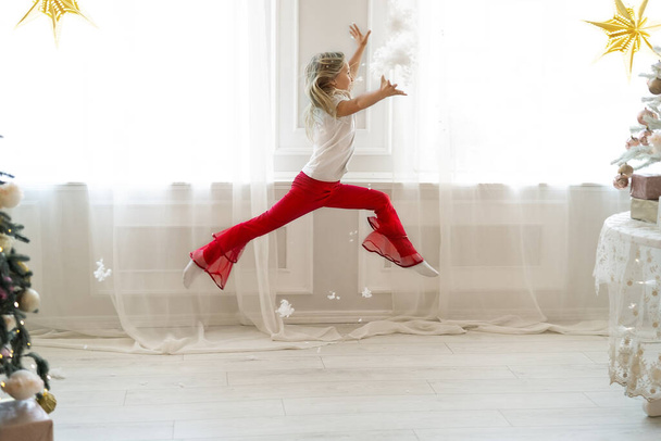 Cute adorable little blond caucasian kid girl fun make dramatic ballet leap contemporary dance in room decor with xmas holiday tree against big window on background. Funny bell pants hipster style - Foto, Imagen