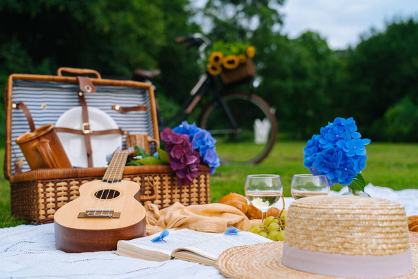 Letní piknik za slunečného dne s chlebem, ovocem, kyticemi, hortenzií, skleničkovým vínem, slamákem, knihou a ukulele. Piknikový koš na trávě s jídlem a pitím na bílé pletenině. Selektivní zaměření - Fotografie, Obrázek