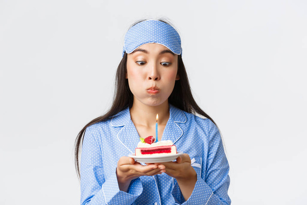 Celebration, lifestyle and holiday concept. Close-up of funny cute asian girl in sleeping mask and pajama being congratulated with birthday in bed, blowing candle on cake and making wish - Photo, Image