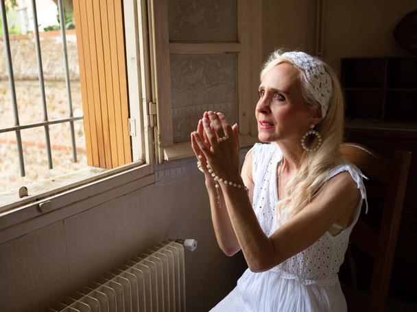 Horizontales Porträt einer reifen Frau, die mit einem Rosenkranz am Fenster betet - Foto, Bild