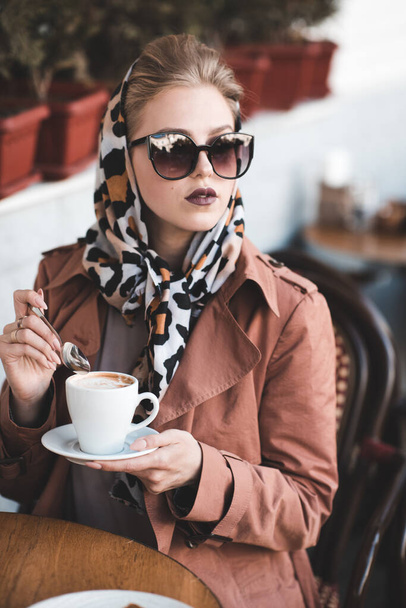 Bella donna 24-25 anni che beve caffè in caffè indossando giacca alla moda, occhiali da sole e sciarpa di seta all'aperto.  - Foto, immagini