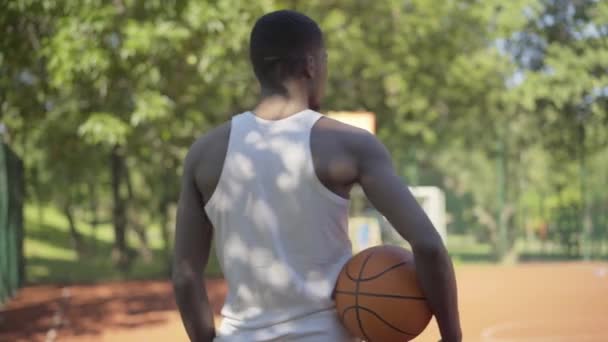 Back view of African American sportsman in white shirt standing with ball on sunny basketball court, turning to camera and smiling. Young confident handsome man posing outdoors on sunny day. - Πλάνα, βίντεο
