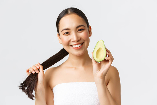 Beauty, personal care, spa and skincare concept. Close-up of happy good-looking asian woman in bath towel, showing hair and avocado, promo of shampoo, haircare masks or conditioner - 写真・画像