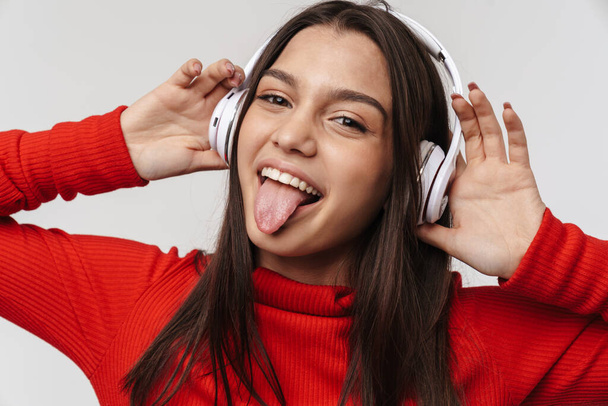 Photo of happy caucasian woman sticking out her tongue while using headphones isolated over white wall - Foto, imagen