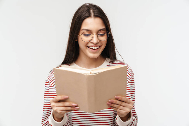Photo of happy brunette young woman wearing eyeglasses smiling and reading book isolated over white background - Фото, изображение