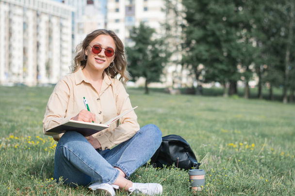 молодая кавказская девушка сидит на траве с книгой и ручкой в кампусе - Фото, изображение