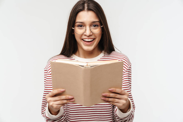 Фото щасливої брюнетки молода жінка в окулярах посміхається і читає книгу ізольовано на білому тлі
 - Фото, зображення
