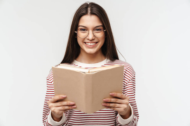 Photo of happy brunette young woman wearing eyeglasses smiling and reading book isolated over white background - Photo, Image
