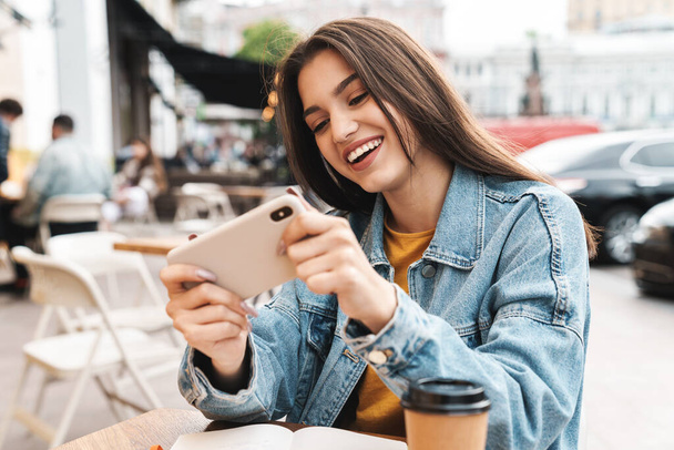 Kuva iloinen brunette nainen pelaa videopeliä matkapuhelimella istuessaan kadulla kahvilassa ulkona - Valokuva, kuva