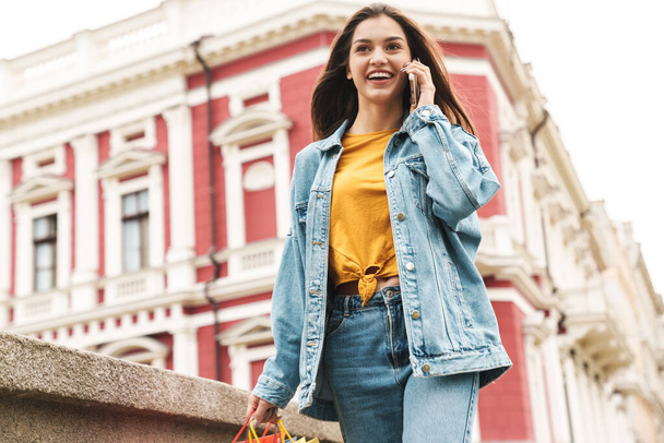 Image of young happy woman in denim wear talking on cellphone while walking with shopping bags on city street - Zdjęcie, obraz