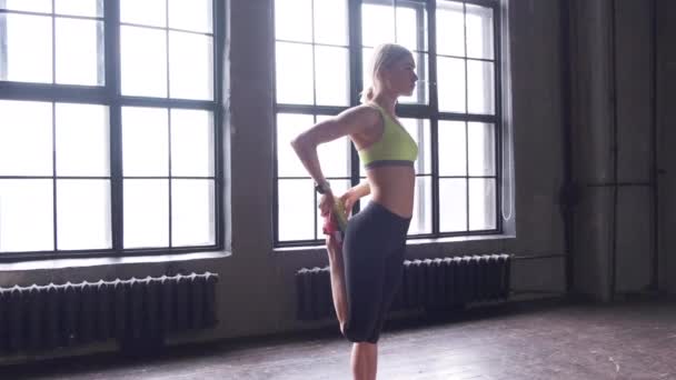 Vista lateral uma jovem mulher loira atlética fazendo um aquecimento alongamento em pé em um tapete em um estúdio de fitness com grandes janelas
 - Filmagem, Vídeo