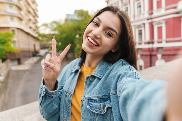 Image of attractive happy woman taking selfie photo on cellphone and gesturing peace sign on city street - Fotoğraf, Görsel