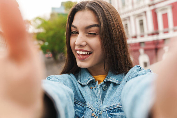 Image of attractive happy woman taking selfie photo on cellphone and winking on city street - Fotó, kép