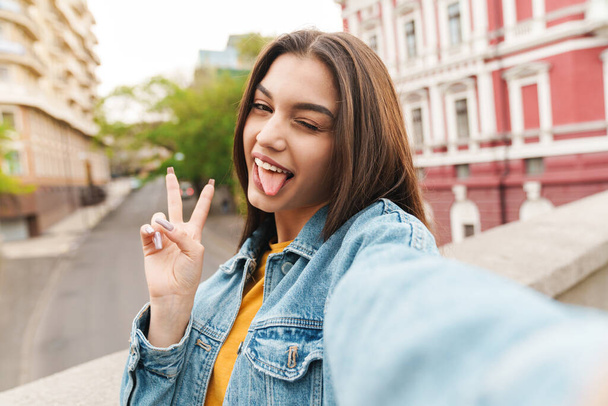 Image of amusing happy woman taking selfie photo on cellphone and gesturing peace sign on city street - Φωτογραφία, εικόνα