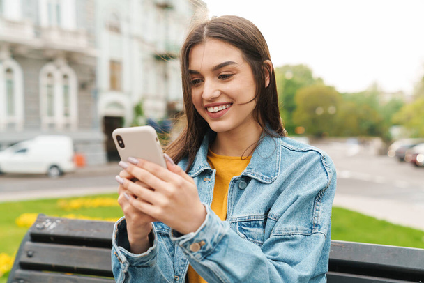Image of attractive happy woman smiling and using mobile phone while sitting on bench at city street - Фото, изображение