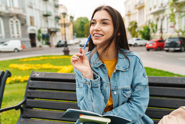 Image of attractive happy woman smiling and making notes in diary while sitting on bench at city street - Фото, зображення