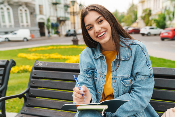 Image of attractive happy woman smiling and making notes in diary while sitting on bench at city street - Foto, Imagen