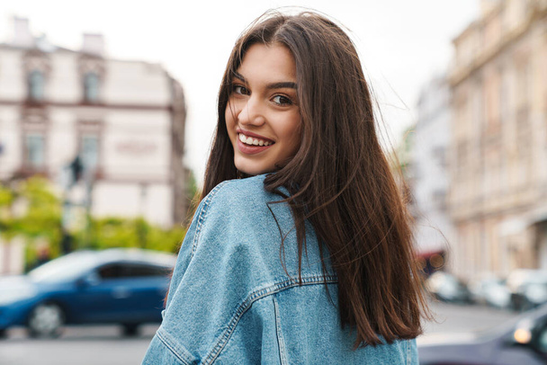 Image of attractive happy woman in denim jacket smiling at camera while walking on city street - Foto, Bild