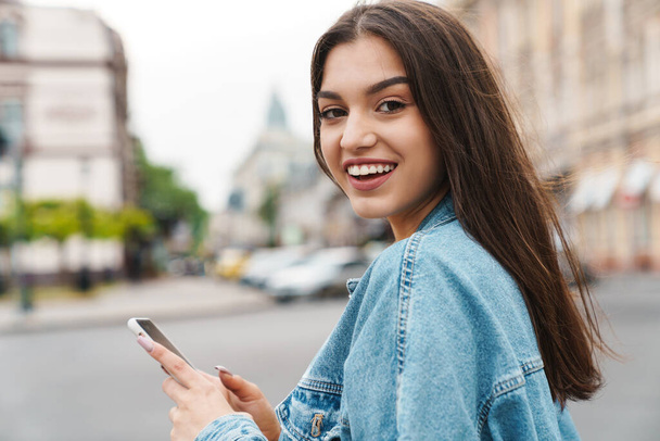 Image of attractive happy woman in denim jacket smiling and using smartphone while walking on city street - Fotó, kép