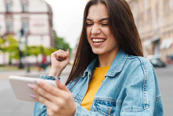 Image of attractive excited woman playing video game on cellphone while walking on city street - Фото, изображение