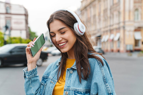 Image of nice laughing woman listening music with smartphone and wireless headphones on city street - Фото, изображение