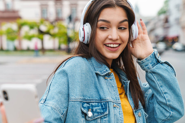 Image of nice laughing woman listening music with smartphone and wireless headphones on city street - 写真・画像