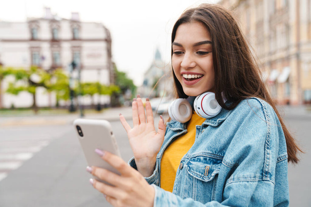 Image of charming cheerful woman with wireless headphones using mobile phone on city street - Фото, зображення