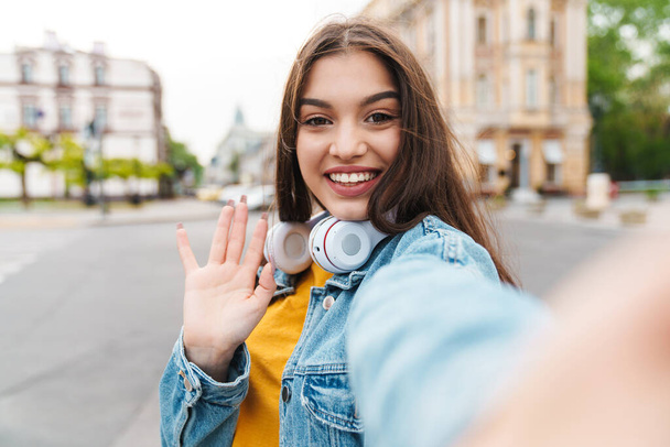 Image of cheerful woman with headphones waving hand and taking selfie on cellphone on city street - Foto, Imagen