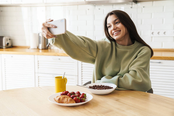 Photo of smiling nice woman taking selfie photo on cellphone while having breakfast in modern kitchen - Фото, зображення