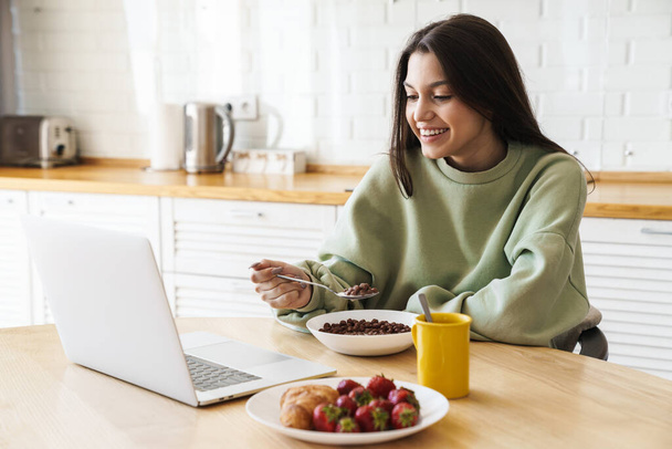 Photo of cheerful beautiful woman using laptop while having breakfast in modern kitchen - Zdjęcie, obraz