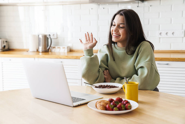 Photo of cheerful beautiful woman waving hand and using laptop while having breakfast in modern kitchen - Фото, зображення
