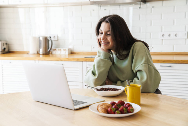 Photo of cheerful beautiful woman using laptop while having breakfast in modern kitchen - Fotografie, Obrázek