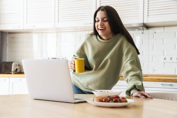 Photo of cheerful beautiful woman using laptop while having breakfast in modern kitchen - Φωτογραφία, εικόνα