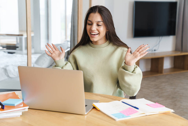 Photo of cheerful student woman waving hands and using laptop while doing homework in living room - Фото, изображение
