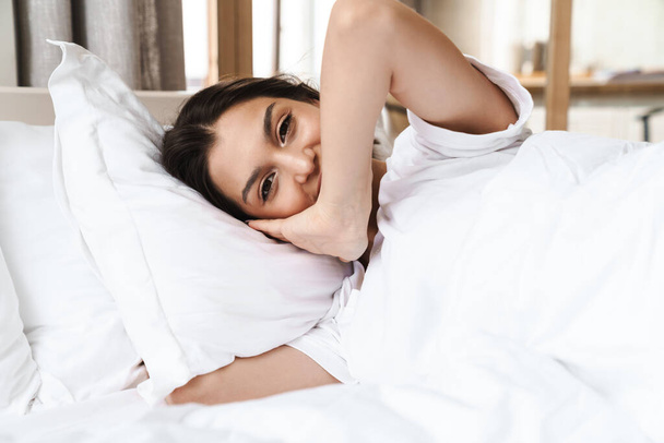 Photo of pretty brunette woman in basic t-shirt smiling while lying on bed after sleep or nap - Zdjęcie, obraz