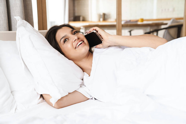 Photo of pretty happy woman smiling and talking on smartphone while lying in bed on white pillow after sleep or nap - Photo, Image