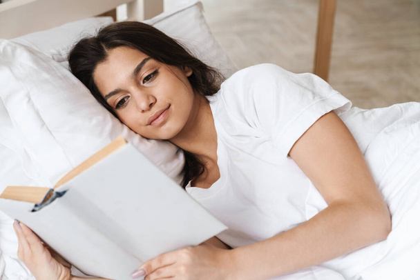 Photo of focused pleased beautiful woman reading book while lying on bed after sleep or nap - Φωτογραφία, εικόνα