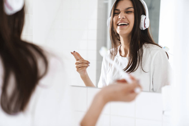 Photo of laughing woman pointing finger at mirror while using wireless headphone and cleaning teethes in bathroom - Photo, Image
