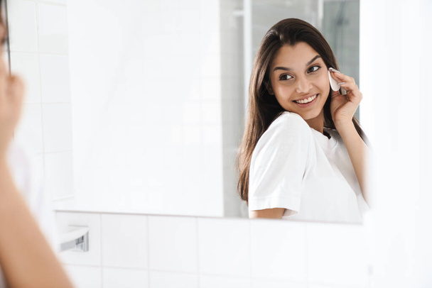 Zdjęcie szczęśliwej miłej kobiety uśmiechającej się i czyszczącej twarz z podkładką bawełnianą patrząc na lustro w łazience - Zdjęcie, obraz
