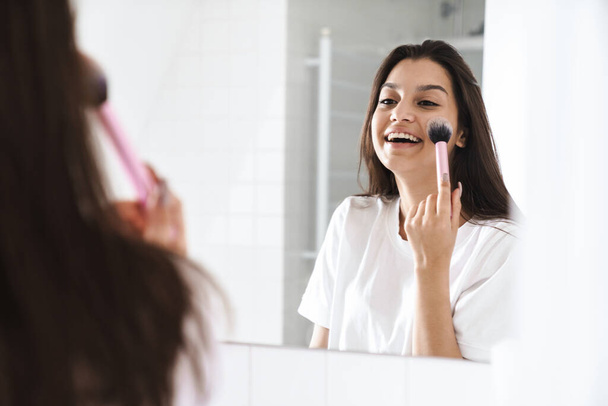 Photo of happy nice woman smiling and using powder brush while looking at mirror in bathroom - Valokuva, kuva