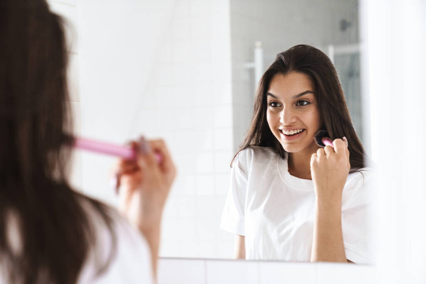 Photo of happy nice woman smiling and using powder brush while looking at mirror in bathroom - Foto, imagen