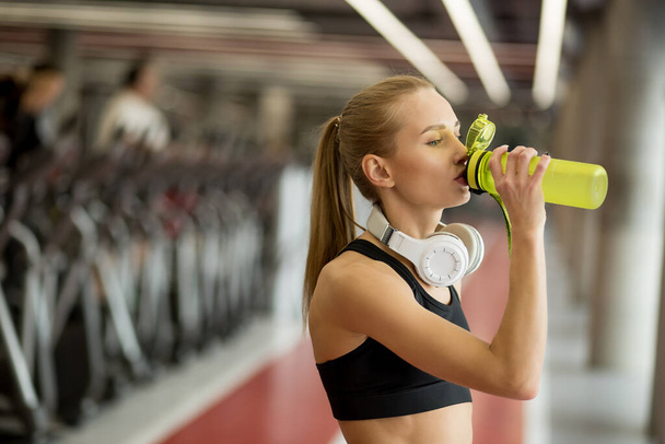 blond athletic woman wearing black closes drinking water in gym - Фото, зображення