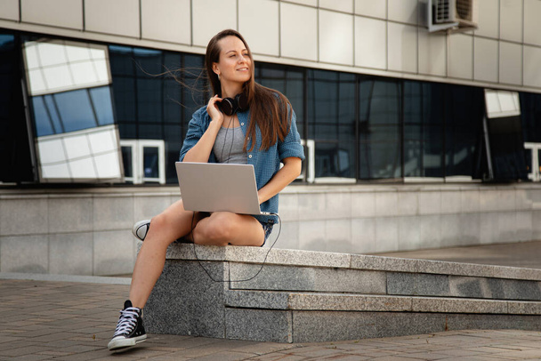 young beautiful girl sit at work at the laptop and listen to music while sitting on the parapet - Photo, image