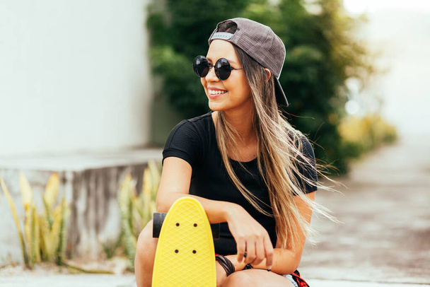 Young skateboarder wearing sunglasses and cap sitting on the sidewalk - Zdjęcie, obraz