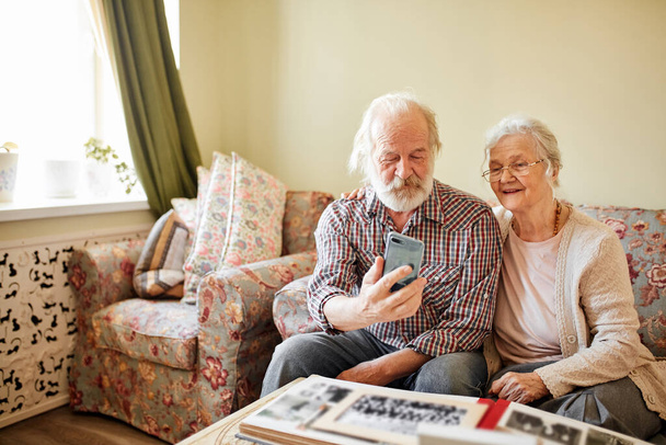 Happy senior couple with smartphone taking selfie at home, old age, technology. - Foto, afbeelding