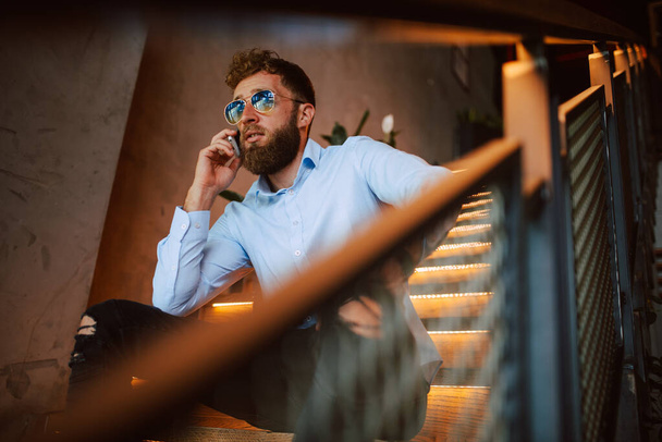 Młody biały mężczyzna w okularach przeciwsłonecznych siedzący na schodach w kawiarni rozmawia przez telefon. Przystojny biznesmen w restauracji - Zdjęcie, obraz