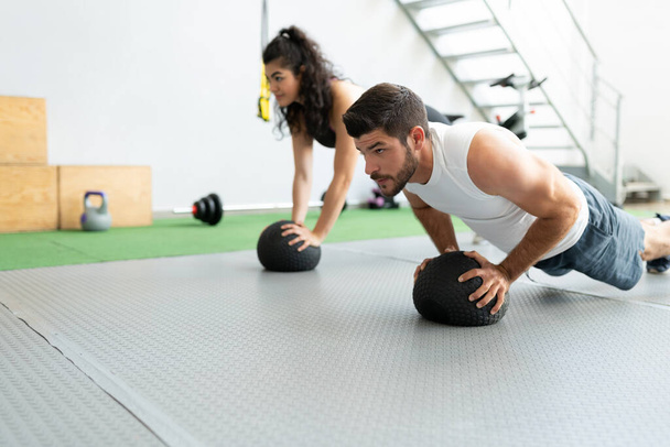 Toegewijd Latijns-Amerikaans jong paar doet push-ups op medicijnballen in fitness club - Foto, afbeelding