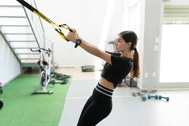 Attractive Hispanic young woman exercising with TRX band at fitness club - Fotografie, Obrázek
