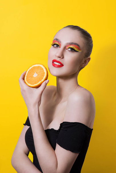 Fashion girl with bright makeup, red lipstick, a bunch on her head, holds a ripe orange on a yellow background. She smiles, surprised, bites, licks the fruit. Close up portrait, copy space, mock up - Foto, immagini