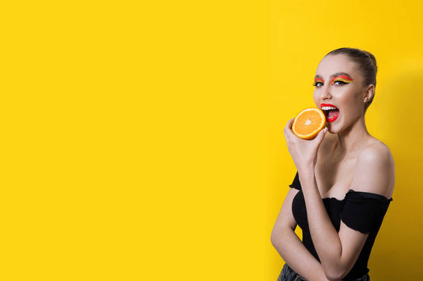 Fashion girl with bright makeup, red lipstick, a bunch on her head, holds a ripe orange on a yellow background. She smiles, surprised, bites, licks the fruit. Close up portrait, copy space, mock up - Foto, Imagem